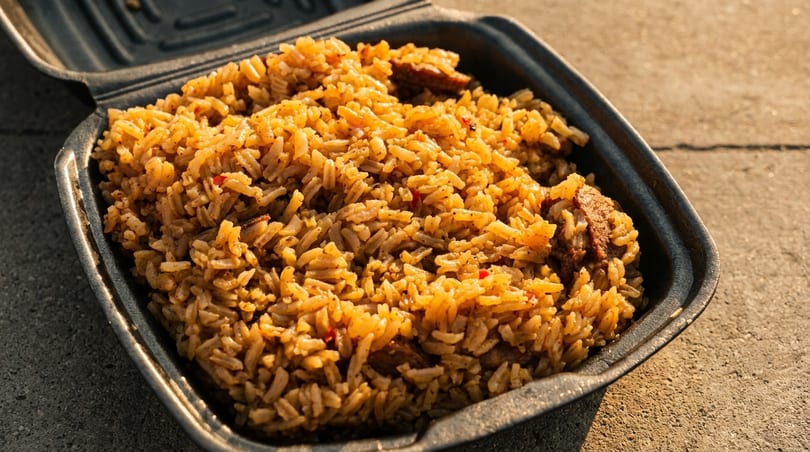 Close-up of seasoned rice with meat pieces in a black takeout container on a concrete surface.