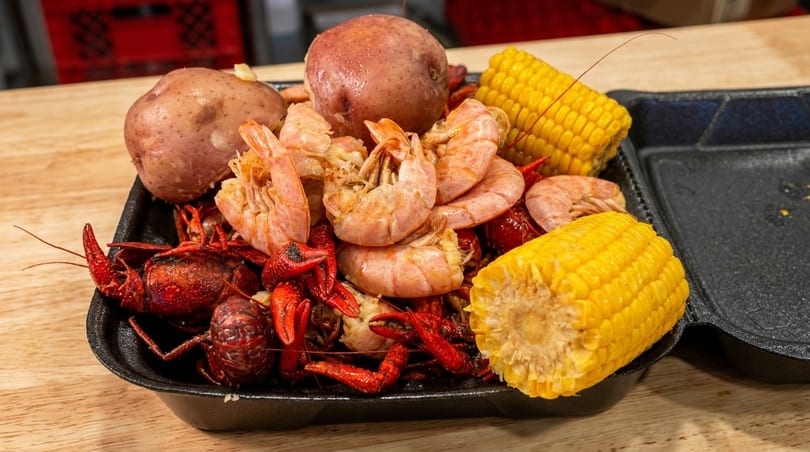 Boiled crawfish, shrimp, potatoes, and corn on the cob in a black takeout container.
