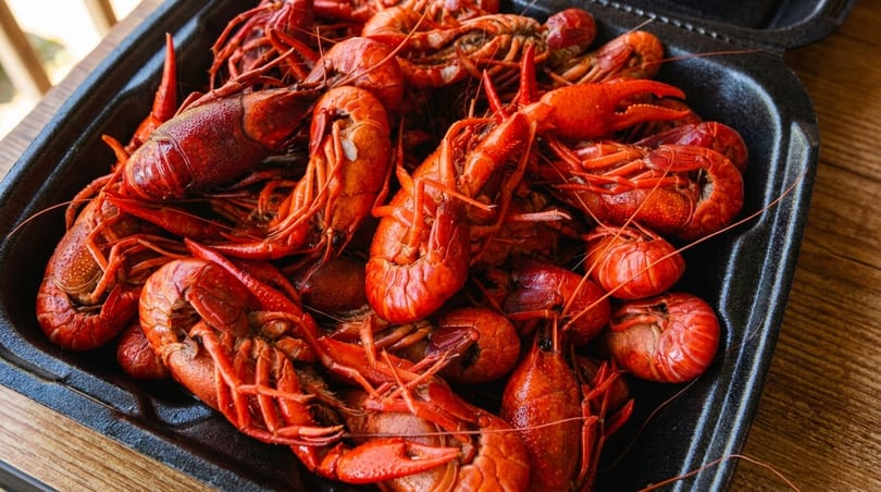 A black takeout container overflows with bright red, cooked crawfish on a wooden table.